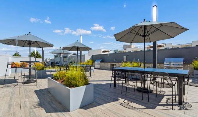 Three different seating areas with umbrellas surrounded by lush landscaped planter boxes and a clear blue sky with some clouds.