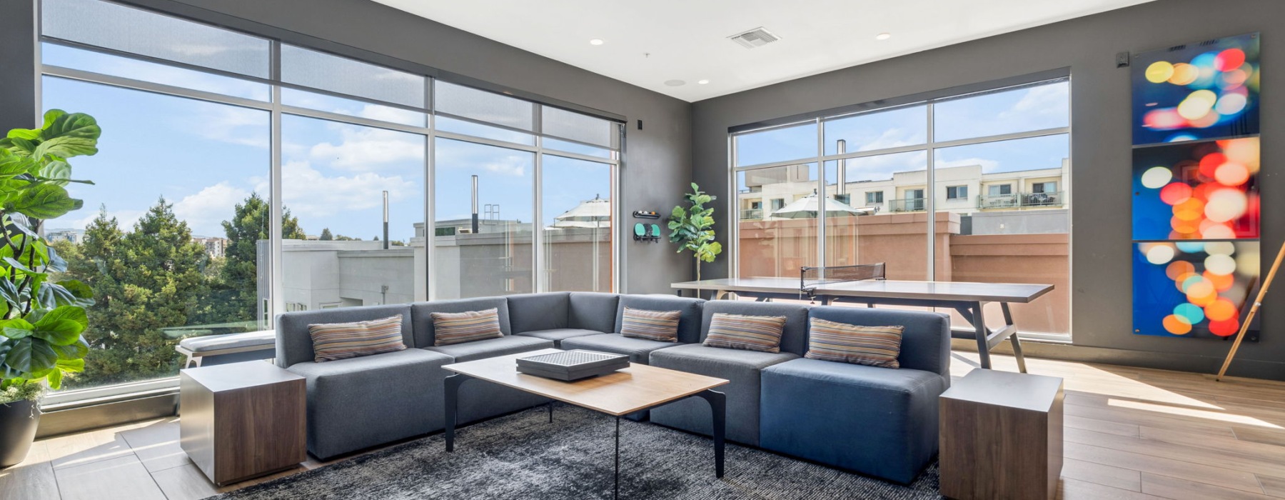 Large Room with huge, blue l-shaed modular sofa , coffee table, two sets of large oversized windows and a ping-pong table.